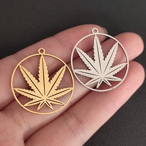 May include: Two circular pendants with a cannabis leaf design. One is gold-coloured, the other silver-coloured. Each pendant has a circular frame around the leaf, with a small loop at the top for attaching to a chain or other jewellery.