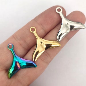 May include: Three metal whale tail charms in gold, silver, and iridescent rainbow colors. Each charm has a small loop at the top for attaching to jewelry or other crafts. The charms are shiny and reflect light.
