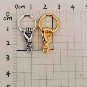 K&ouml;nnte beinhalten: Zwei Metall-Hand-Anh&auml;nger, einer silber und einer gold, mit einem Ring oben. Die H&auml;nde sind in Faustform mit dem Zeigefinger nach oben.
