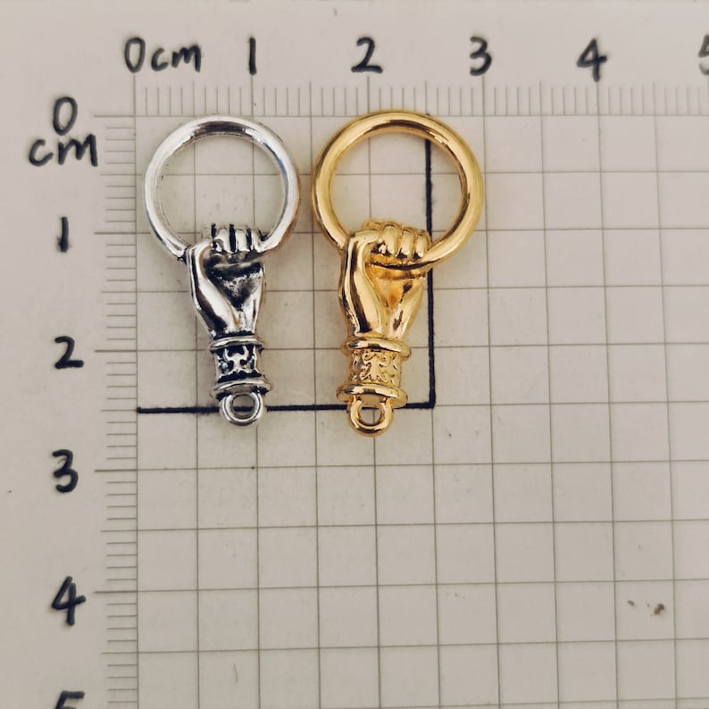 K&ouml;nnte beinhalten: Zwei Metall-Hand-Anh&auml;nger, einer silber und einer gold, mit einem Ring oben. Die H&auml;nde sind in Faustform mit dem Zeigefinger nach oben.