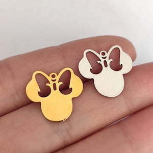 May include: Two metal charms in the shape of a bow and head. One charm is gold-colored, the other is silver-colored. The charms have a small hole at the top for stringing. The charms are on a light background.