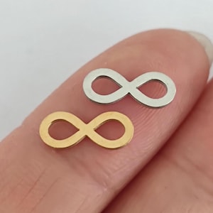 Könnte beinhalten: Zwei Unendlichkeitszeichen-Anhänger, einer goldfarben und einer silberfarben, werden gezeigt. Die kleinen Anhänger sind wahrscheinlich für Schmuck oder Bastelarbeiten gedacht. Die Symbole sind vor einem weichen, verschwommenen Hintergrund platziert.