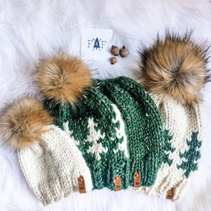 May include: Three knitted winter hats with faux fur pom-poms. Two hats are cream-colored with white snowflake patterns, and one is green with a white snowflake pattern. Each hat has a small leather tag with the letters "FAF".