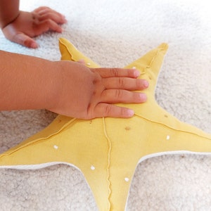 May include: A yellow fabric starfish with white stitching and white dots. The starfish is lying on a white fuzzy surface.