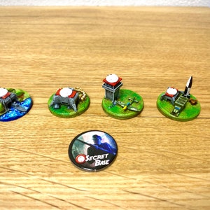 Könnte beinhalten: Fünf Miniatur-Militärbasen mit Radaranlagen, einem Flugzeug und einer Rakete. Die Basen stehen auf grünen und blauen runden Sockeln. Ein runder Knopf mit dem Text "Geheime Basis" befindet sich in der Mitte des Bildes.