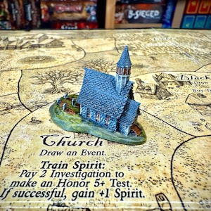 Könnte beinhalten: Eine blaue Miniaturkirche mit Turm, auf einem grünen Sockel, liegt auf einem Spielbrett. Das Brett zeigt eine Karte mit dem Text "Kirche" und Spielanweisungen. Das Kirchenmodell ist ein Spielstein.