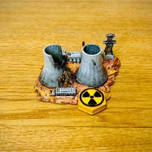 Puede incluir: Un modelo en miniatura de una planta de energía nuclear con dos torres de refrigeración, un pequeño edificio y una línea eléctrica. El modelo está colocado sobre una base marrón con una señal de advertencia de radiación amarilla.