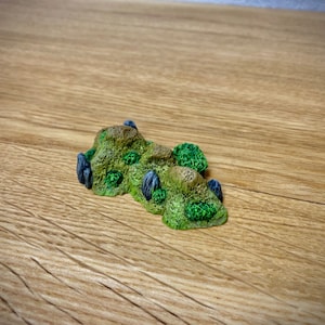 May include: A miniature green and brown terrain model with small black rocks, perfect for tabletop gaming or miniature painting.