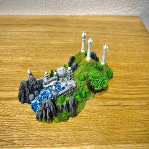 Puede incluir: Un modelo en miniatura de una ciudad de fantasía con torres blancas, un edificio de piedra y un río azul. La ciudad está rodeada de árboles verdes y rocas, y está colocada sobre una superficie de madera marrón.