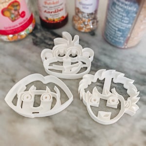 May include: Three white plastic cookie cutters shaped like cartoon faces. The cutters are on a white marble surface. The cutters are for making cookies.