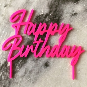 Op de afbeelding: Een roze acryl taarttopper met de woorden "Happy Birthday" in een cursief schrift.