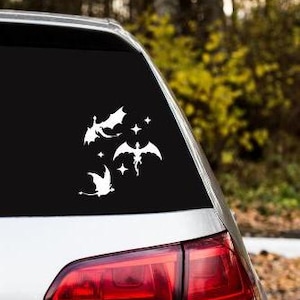 May include: White vinyl car decal featuring three stylized dragon silhouettes and several stars. The dragons have outstretched wings and are in various poses. The decal is applied to the black rear window of a car.