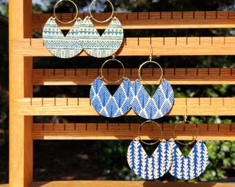 May include: Three pairs of blue and white patterned earrings on a wooden display stand. The earrings are made of cork and have gold hoops.
