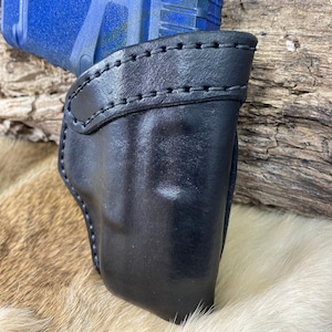 Può includere: Un fodero in pelle nera per una pistola blu. Il fodero ha un bordo cucito ed è progettato per adattarsi perfettamente alla pistola.