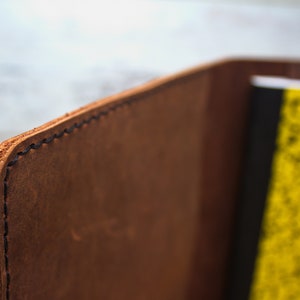 K&ouml;nnte beinhalten: Nahaufnahme der Kante eines braunen Leder-Notizbuchdeckels mit schwarzen N&auml;hten. Der Deckel ist &uuml;ber ein gelbes Notizbuch mit schwarzen Linien gefaltet.