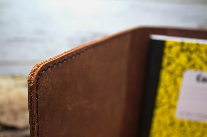 K&ouml;nnte beinhalten: Nahaufnahme der Kante eines braunen Leder-Notizbuchdeckels mit schwarzen N&auml;hten. Der Deckel ist &uuml;ber ein gelbes Notizbuch mit schwarzen Linien gefaltet.