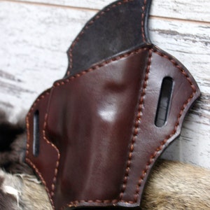 May include: A brown leather holster with a black leather lining and brown stitching. The holster has two belt loops on the back.