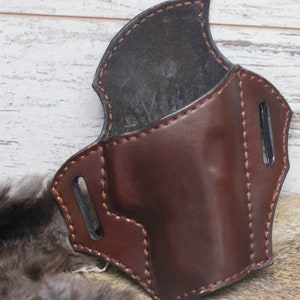 May include: A brown leather holster with a black leather lining. The holster has a stitched edge and two slots on the side.
