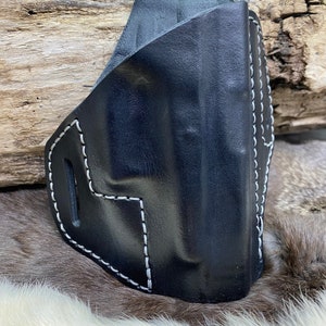 Può includere: Una fondina in pelle nera con cuciture bianche. La fondina ha una forma curva e una fessura sul lato. Le cuciture sono visibili lungo i bordi e le cuciture. Lo sfondo è un pezzo di legno e pelliccia.