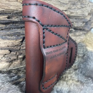 May include: A brown leather holster with black stitching and the text "SNAKE RIVER HOLSTERS GLOCK 19 SRA225".