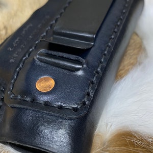 Peut inclure: &Eacute;tui en cuir noir avec un clip en m&eacute;tal et un rivet en cuivre. L'&eacute;tui est pli&eacute; et pr&eacute;sente une couture cousue.