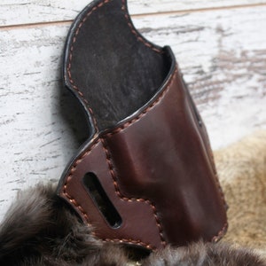 May include: A brown leather holster with stitching detail. The holster has a flap that covers the top of the opening.
