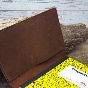 K&ouml;nnte beinhalten: Ein braunes Leder-Notizbuchcover mit einem gen&auml;hten Rand. Das Cover ist ge&ouml;ffnet und zeigt ein gelbes Kompositions-Notizbuch mit schwarzer Schrift auf der Vorderseite.