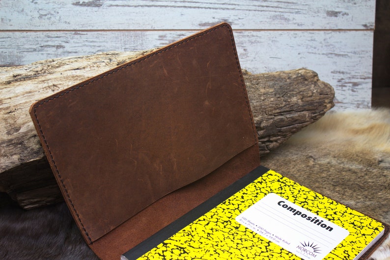 K&ouml;nnte beinhalten: Ein braunes Leder-Notizbuchcover mit einem gen&auml;hten Rand. Das Cover ist ge&ouml;ffnet und zeigt ein gelbes Kompositions-Notizbuch mit schwarzer Schrift auf der Vorderseite.