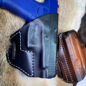 Puede incluir: Una funda de cuero negra con costuras blancas para una pistola azul. La funda tiene un bucle para el cinturón y una segunda funda en el lateral.