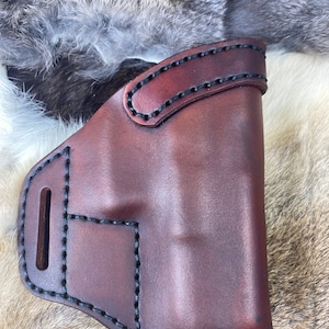 Puede incluir: Una funda de cuero marrón con costuras negras. La funda tiene un bucle para el cinturón y una solapa que cubre la abertura.