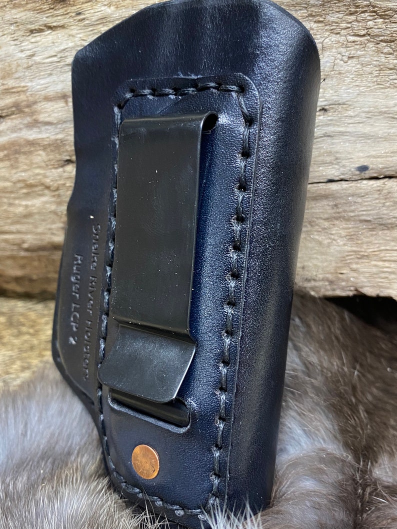Peut inclure: Un &eacute;tui en cuir bleu fonc&eacute; avec une pince m&eacute;tallique et des coutures. L'&eacute;tui est con&ccedil;u pour un pistolet Ruger LCR. "Smith River Holsters" est imprim&eacute; sur le c&ocirc;t&eacute;.
