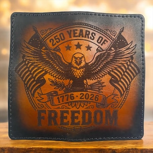 以下が含まれることがあります： 茶色の革製財布。鷲、旗、テキスト「250 YEARS OF FREEDOM 1776-2026」が刻印されています。財布は暗い縁取りと質感のある外観です。愛国的なデザインです。