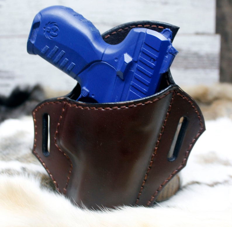 May include: A brown leather holster with a blue handgun inside. The holster has stitching around the edges and two belt loops.