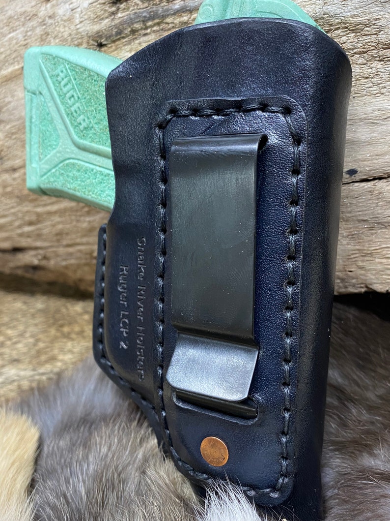 Peut inclure: Un &eacute;tui en cuir noir avec un clip pour un pistolet Ruger LCP 2. L'&eacute;tui est cousu avec du fil blanc et porte une &eacute;tiquette Snake River Holsters.