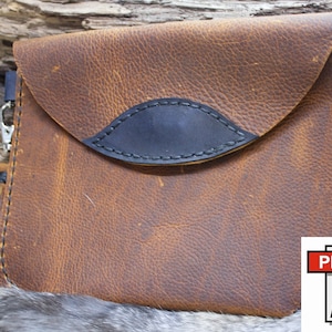 May include: A brown leather clutch with a black accent and a wrist strap. The clutch has a flap closure and a decorative black leather detail. The leather has a distressed look. The clutch is a PDF file.