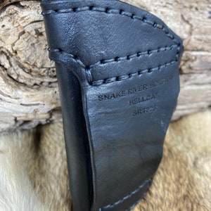 Op de afbeelding: Zwart leren holster voor een Hellcat-pistool. Het holster is gestikt met zwart garen en heeft de tekst "Snake River Holsters Hellcat SRT225" gegraveerd aan de zijkant.