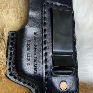 Peut inclure: &Eacute;tui en cuir noir avec un clip en m&eacute;tal et le texte "Snake River Holsters Ruger LCP 2" grav&eacute; sur le c&ocirc;t&eacute;.