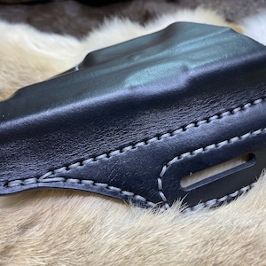 Può includere: Fondina in pelle nera con dettagli di cuciture grigie. La fondina ha un'apertura per una cintura.