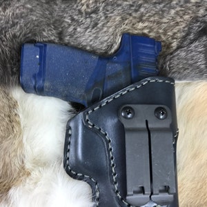 Può includere: Un fodero in pelle nera con una clip grigio scuro per una pistola. Il fodero ha un design cucito ed è mostrato con una pistola blu all'interno.