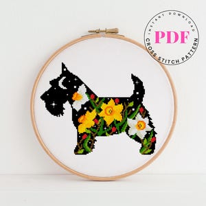 May include: Cross-stitch pattern of a Scottish Terrier dog silhouette. The dog is black with a starry night design on the head and a floral pattern of yellow daffodils and white flowers on the body. The design is in a wooden embroidery hoop. Text reads "INSTANT DOWNLOAD PDF CROSS STITCH PATTERN".