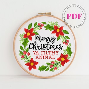 May include: A cross-stitch design in a wooden hoop features a Christmas wreath with red poinsettias, green leaves, and red berries. The text reads "Merry Christmas Ya Filthy Animal" in black and red.