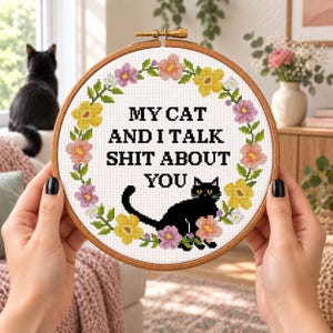 May include: A circular wooden embroidery hoop containing white fabric with a cross-stitch design. The design features a colorful floral wreath surrounding the black text MY CAT AND I TALK SHIT ABOUT YOU and a black cat silhouette at the bottom.