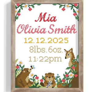 May include: A framed cross-stitch birth announcement with the name "Mia Olivia Smith" in red and pink. The date, weight (8lbs 6oz), and time are also displayed. The design includes woodland animals and floral accents.