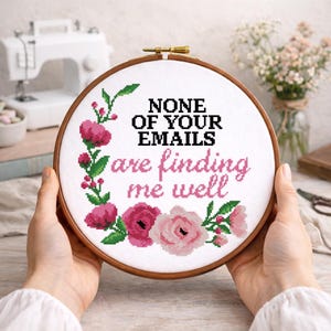 May include: A cross-stitch embroidery hoop with the text "NONE OF YOUR EMAILS are finding me well." The design features pink flowers and green leaves around the text. The hoop is held by hands.