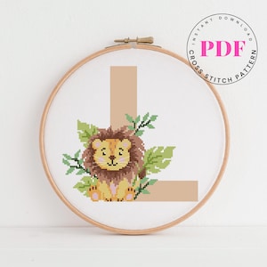 May include: Cross-stitch pattern in a wooden hoop featuring a tan letter "L" with a smiling lion and green leaves. The text "INSTANT DOWNLOAD PDF CROSS STITCH PATTERN" is in a circle around the top right.