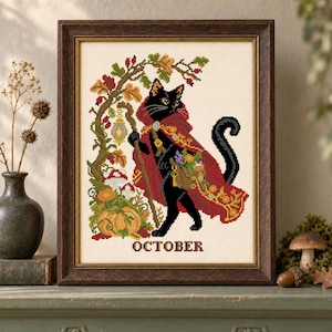 Puede incluir: Un bordado en punto de cruz enmarcado que muestra un gato negro con una capa roja, portando un bastón de madera y un morral. El diseño incluye follaje otoñal, calabazas y setas, con la palabra OCTOBER impresa en marrón en la parte inferior.
