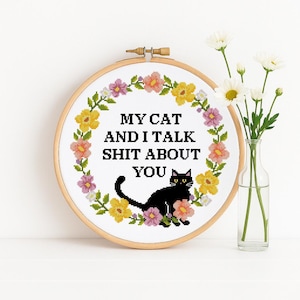 May include: A cross-stitch hoop with the text "MY CAT AND I TALK SHIT ABOUT YOU" in black. The design features a black cat and a floral border with yellow, pink, and green accents. A vase of white flowers is to the right.