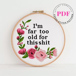 以下が含まれることがあります： 「I'm far too old for this shit」の文字が入ったクロスステッチ刺繍枠。ピンクの花と緑の葉のデザイン。右上隅にある円形のスタンプには「PDF」と「Instant Download Cross Stitch Pattern」と書かれています。