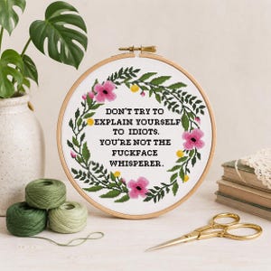 May include: A circular wooden embroidery hoop containing white fabric with a cross-stitch design. The design features a floral wreath in green, pink, and yellow surrounding the black text: DON'T TRY TO EXPLAIN YOURSELF TO IDIOTS. YOU'RE NOT THE FUCKFACE WHISPERER.