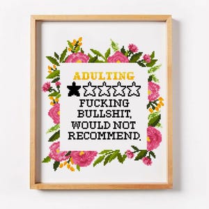 May include: A framed cross-stitch artwork with a wooden frame. The artwork features the word "ADULTING" above a rating of one out of five stars, and the phrase "FUCKING BULLSHIT, WOULD NOT RECOMMEND." Pink flowers and green leaves surround the text.